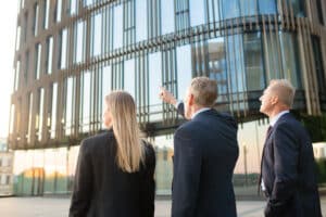 Group of business partners in formal suits pointing at office building, meeting outdoors, discussing real property. Back view. Commercial real estate concept